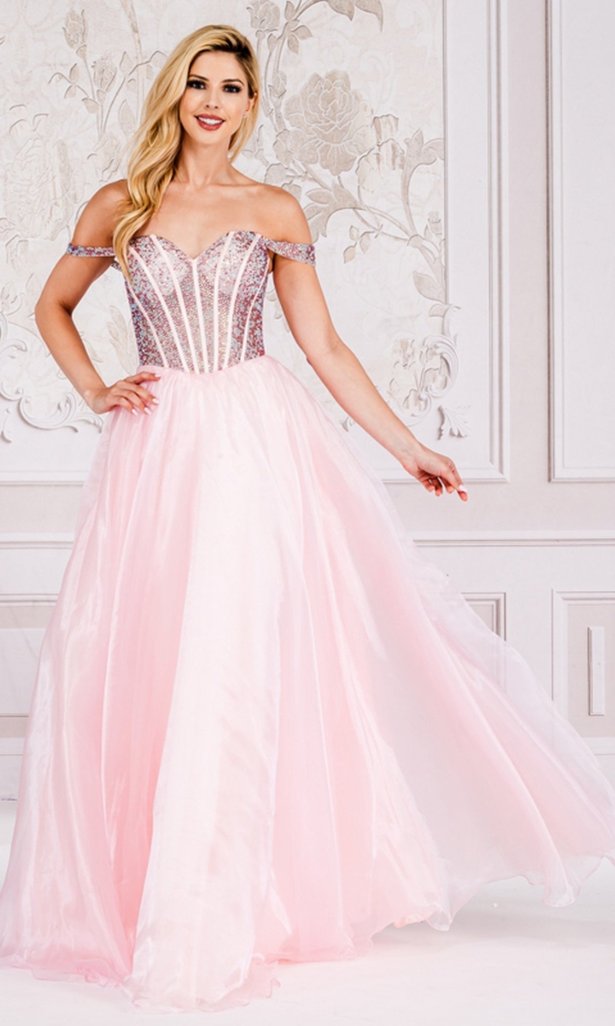 A woman stands indoors wearing the Organza Off-Shoulder Bodice Prom Ball Gown 7040, featuring a fitted, embellished bodice and a flowing skirt.